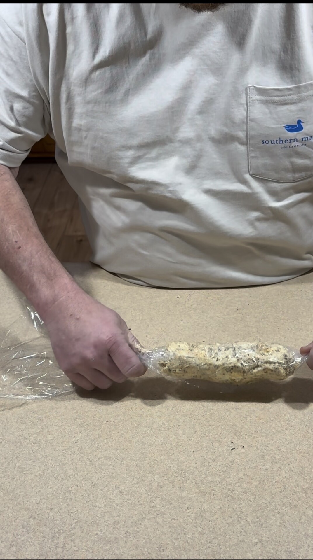 A person in a white shirt is rolling a log-shaped mixture coated in herbs and spices in plastic wrap on a counter.