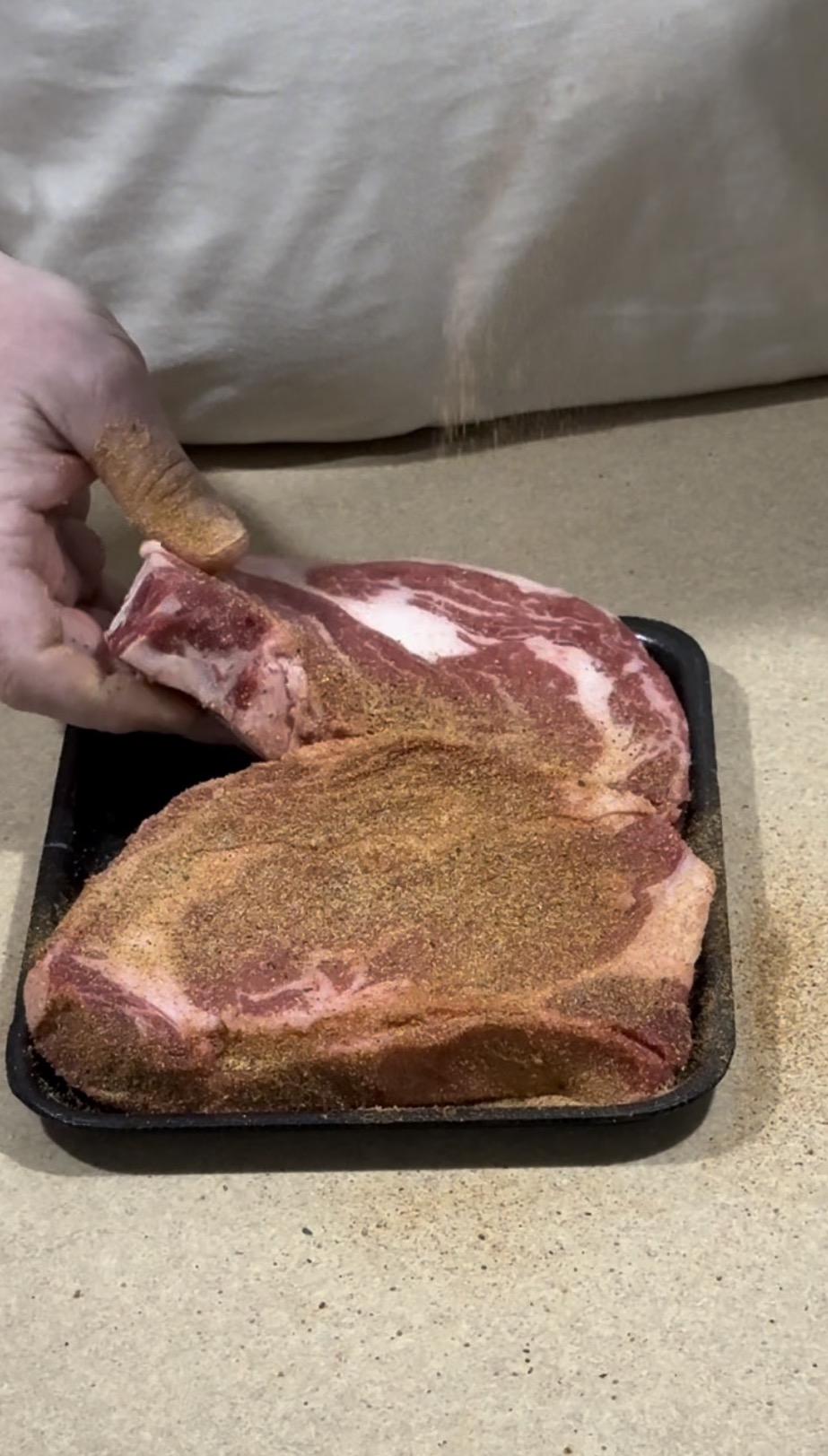 A hand sprinkles seasoning onto two raw bone-in ribeye steaks, one upright, on a black tray placed on a beige countertop.