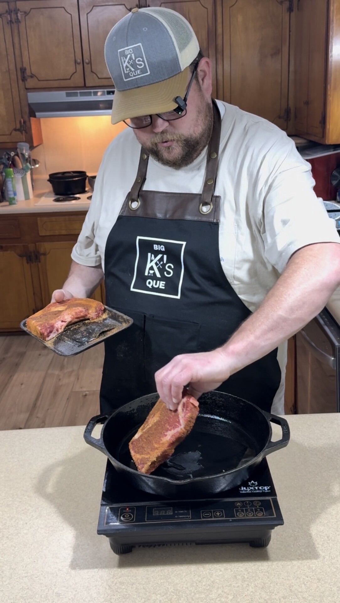 A man wearing a hat and apron places a seasoned steak into a cast iron pan on an induction cooktop in a kitchen with wooden cabinets.