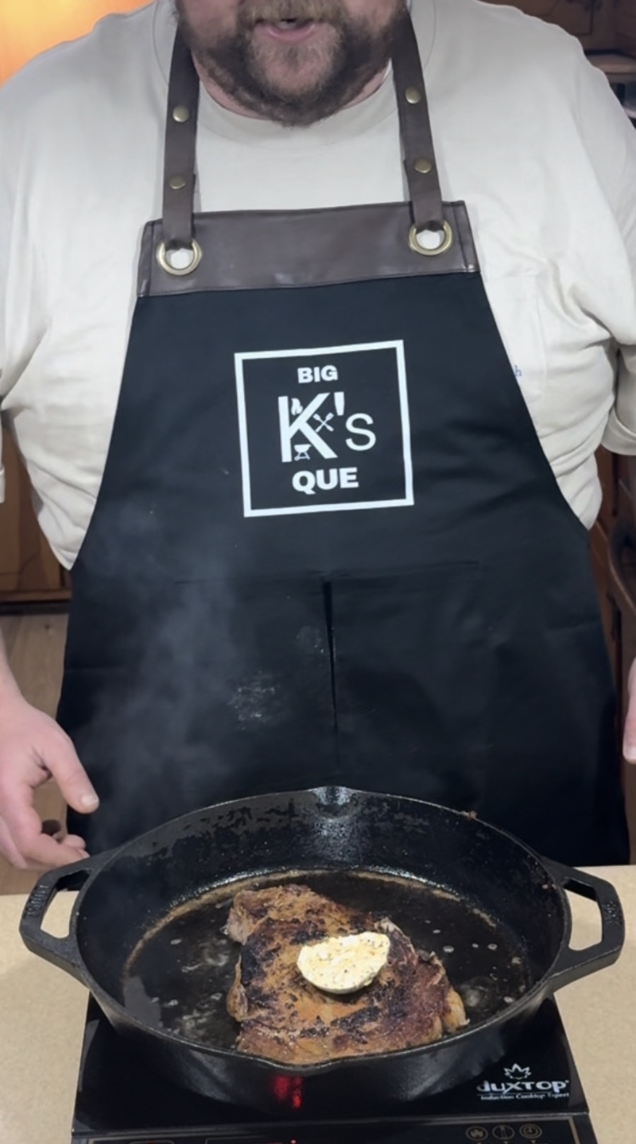 A person wearing a black apron labeled BIG KS QUE cooks a steak in a cast iron skillet. The steak is topped with a pat of melting butter, and steam rises from the pan on a stovetop.