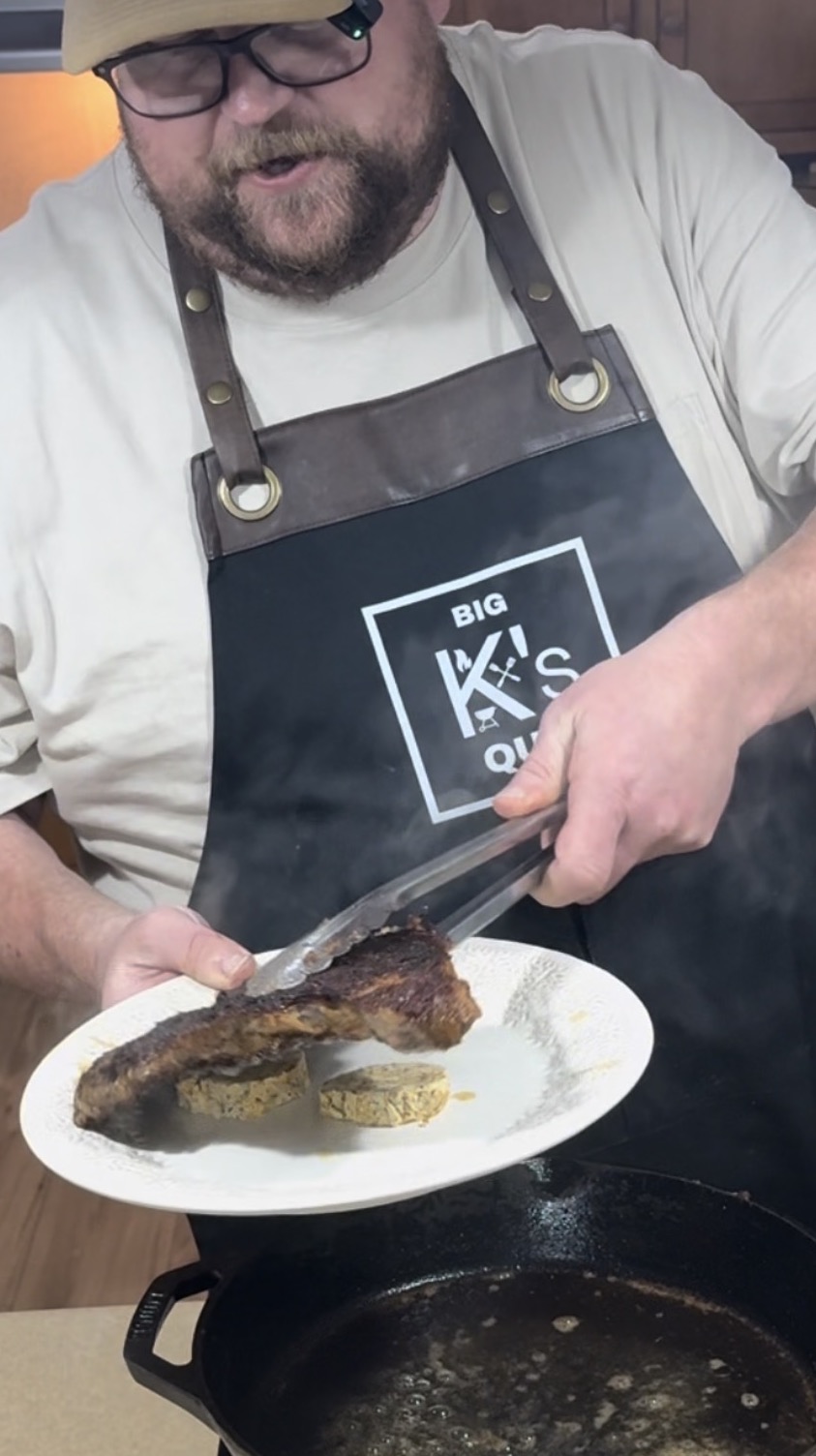 A man wearing glasses and a dark apron uses tongs to transfer a cooked steak onto a white plate with two pieces of herb butter. A cast iron skillet sits on the stove below, with visible steam rising.