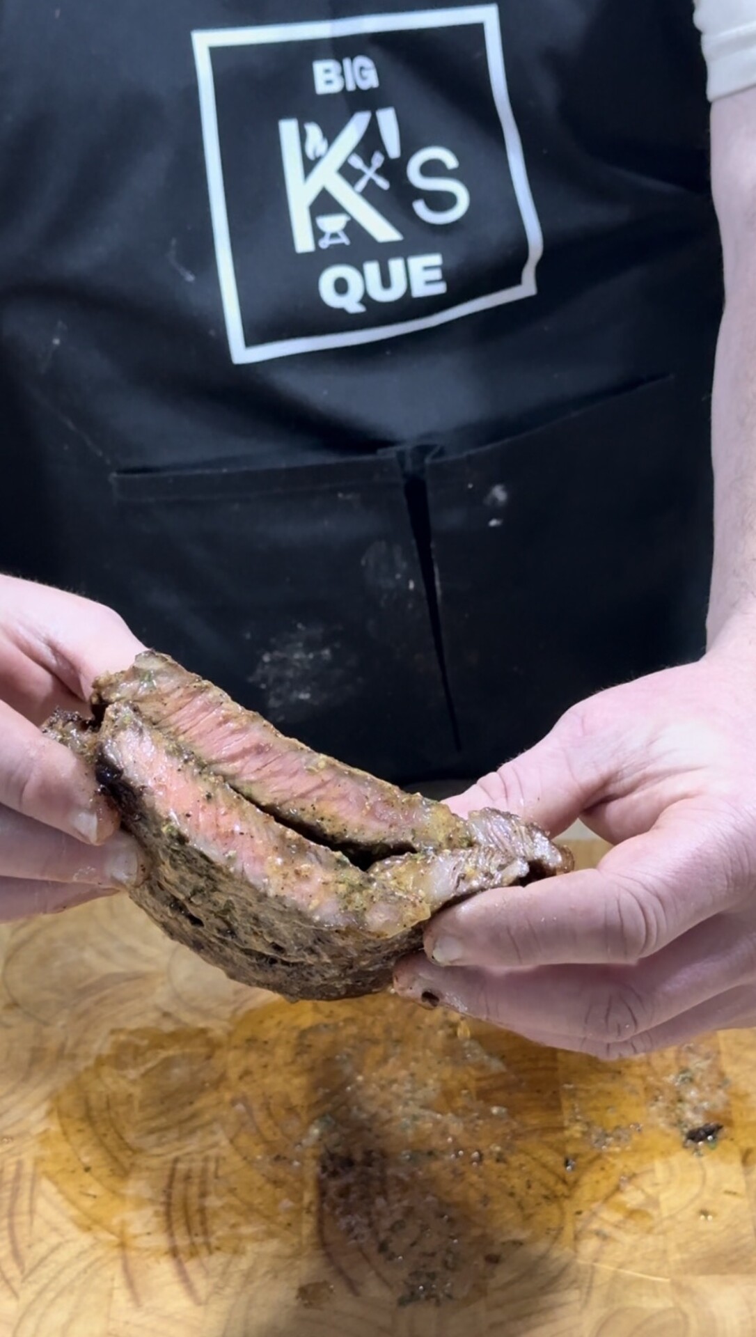 A person wearing a black apron with BIG KS QUE printed on it holds a sliced piece of medium-rare steak over a wooden cutting board.