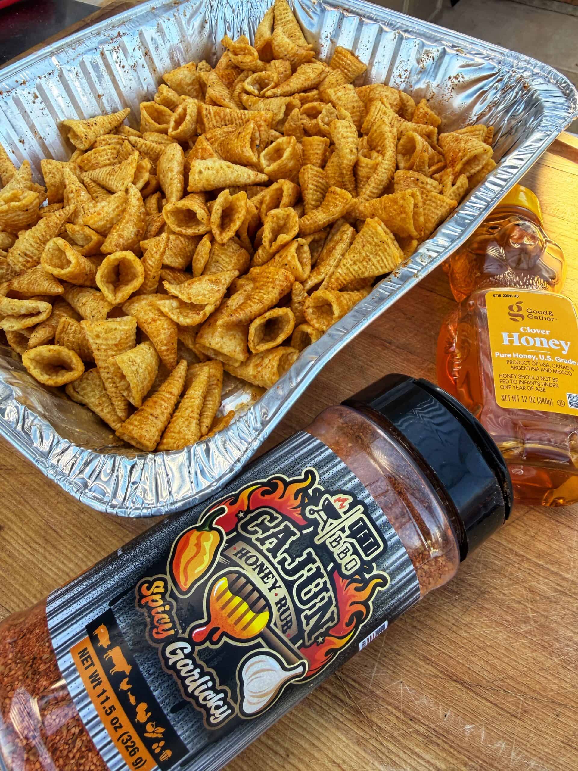 A large foil tray filled with corn snack chips sits on a wooden table, next to a container of Cajun seasoning and a bottle of honey shaped like a bear.