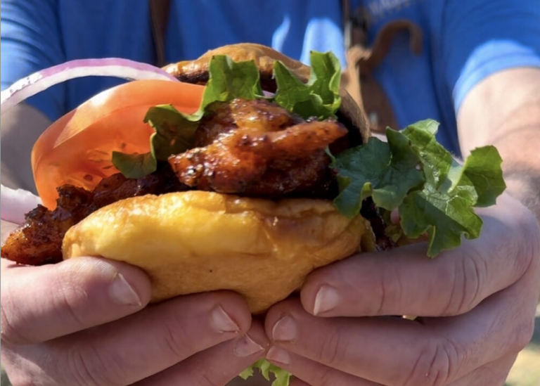 A person holding a large Honey Chipotle Grilled Chicken Sandwich with lettuce, tomato, red onion, and a toasted bun, both hands visible and wearing a blue shirt.