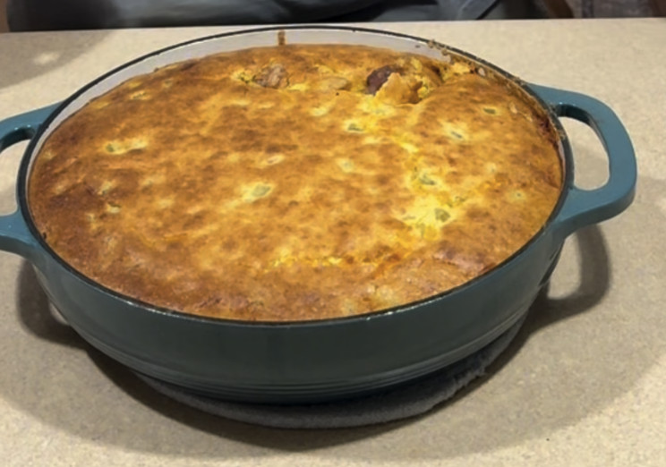 A golden-brown baked cornbread sits in a round, teal-colored Dutch oven on a beige countertop.