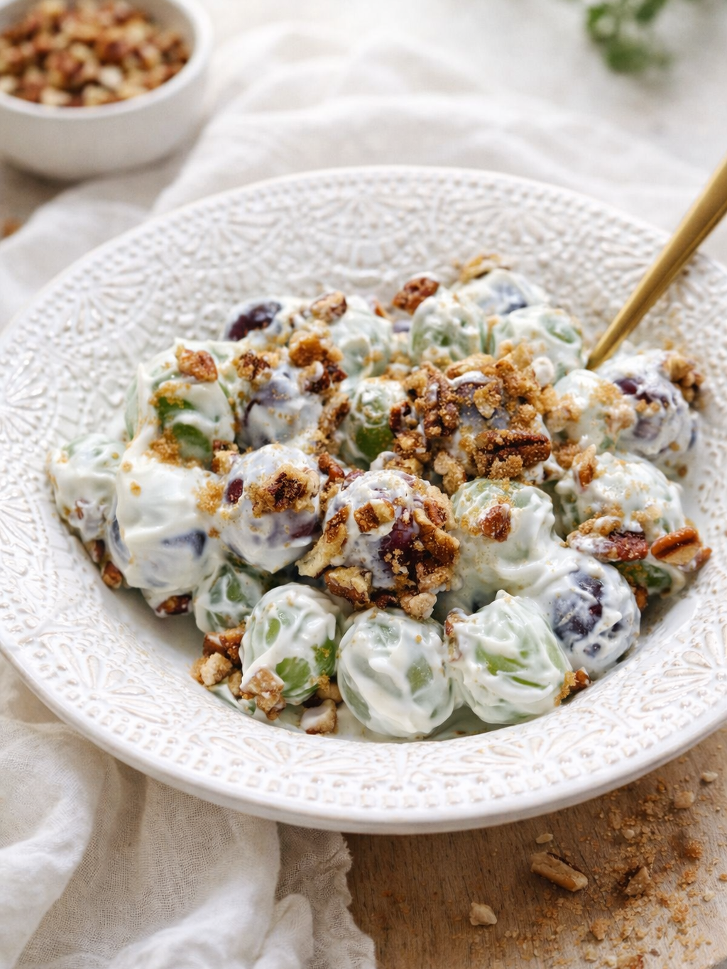 A white textured bowl filled with grape salad coated in creamy dressing, topped with chopped pecans and brown sugar. A gold spoon rests in the salad, and a small dish of extra pecans sits in the background.