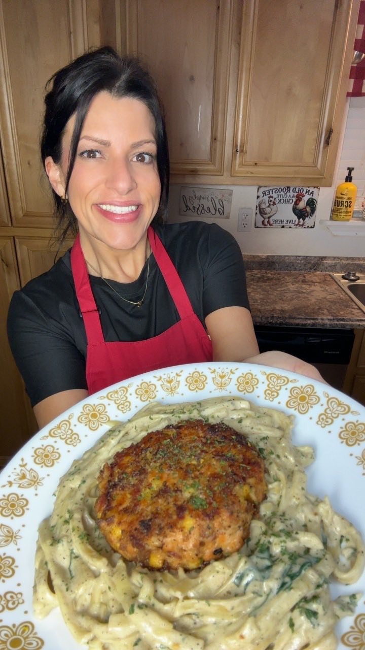 A smiling woman in a red apron holds a plate of creamy pasta topped with a large, seasoned patty. She stands in a kitchen with wooden cabinets and decorative signs in the background.