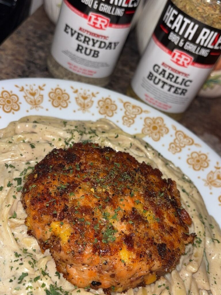 A seasoned crab cake sits atop creamy pasta on a floral plate, garnished with herbs. In the background are bottles of Heath Riles BBQ & Grilling Everyday Rub and Garlic Butter seasoning.