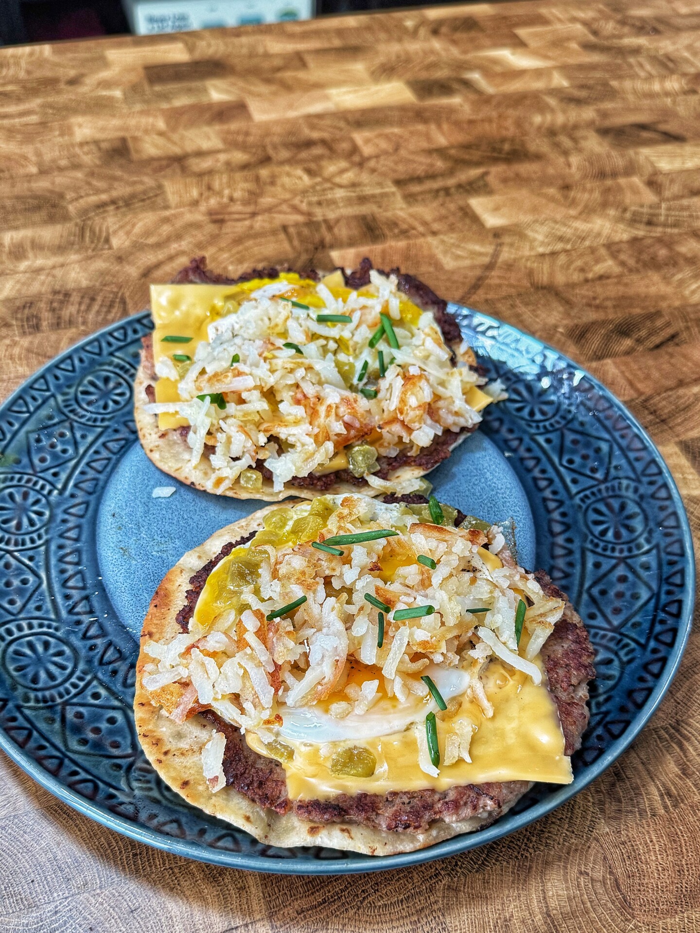 Two open-faced sandwiches on a decorative plate, each topped with a burger patty, a slice of melted cheese, potato sticks, pickles, onions, and chopped chives on a toasted bun, set on a wooden table.