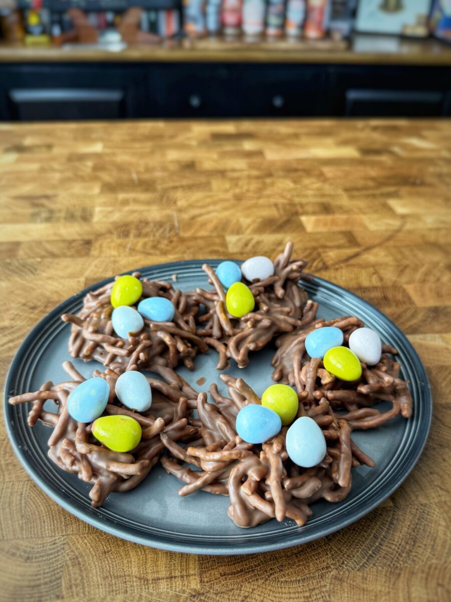 A plate of chocolate-covered chow mein noodle bird nest treats, each topped with pastel-colored candy eggs, sits on a wooden countertop in a kitchen.