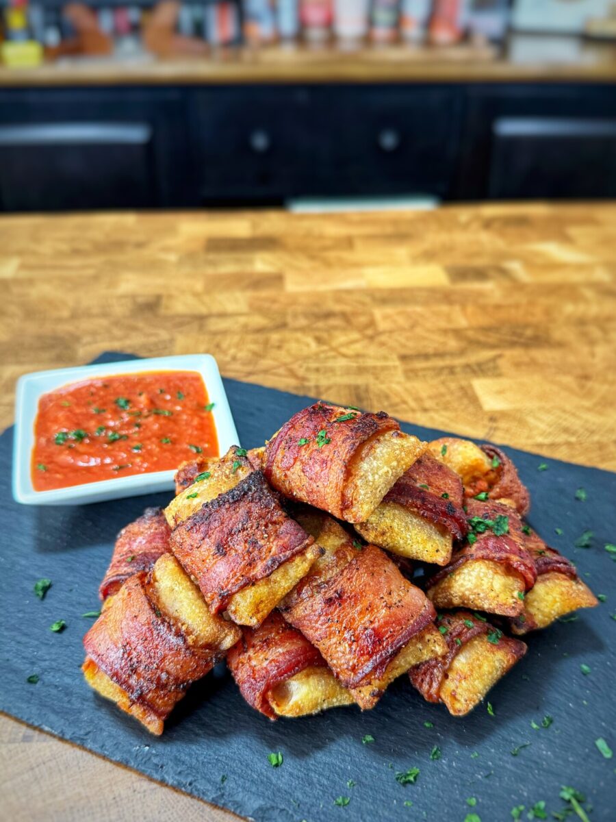 A stack of crispy bacon-wrapped snacks garnished with herbs sits on a slate board, with a small dish of red dipping sauce on the side, on a wooden countertop.