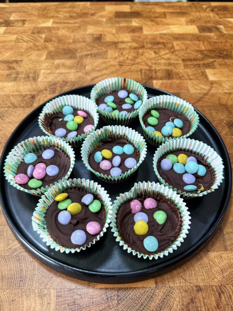 A black plate holds eight cupcake liners filled with chocolate and topped with colorful candy-coated chocolates, arranged on a wooden surface.