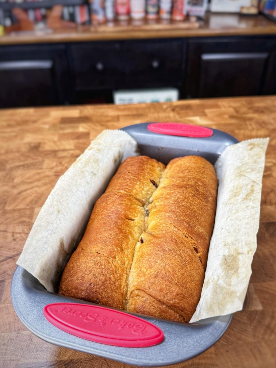 A loaf of freshly baked bread, golden brown on top, rests in a parchment-lined metal baking pan on a wooden countertop with kitchen cabinets and spice jars in the background.