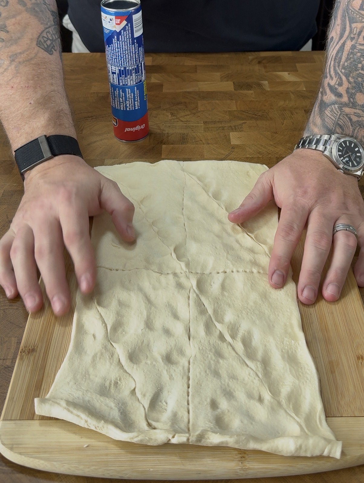 A person’s hands spread out a sheet of dough on a wooden cutting board, with a can of Pillsbury dough in the background on a wooden surface. The person is wearing a watch and a ring.