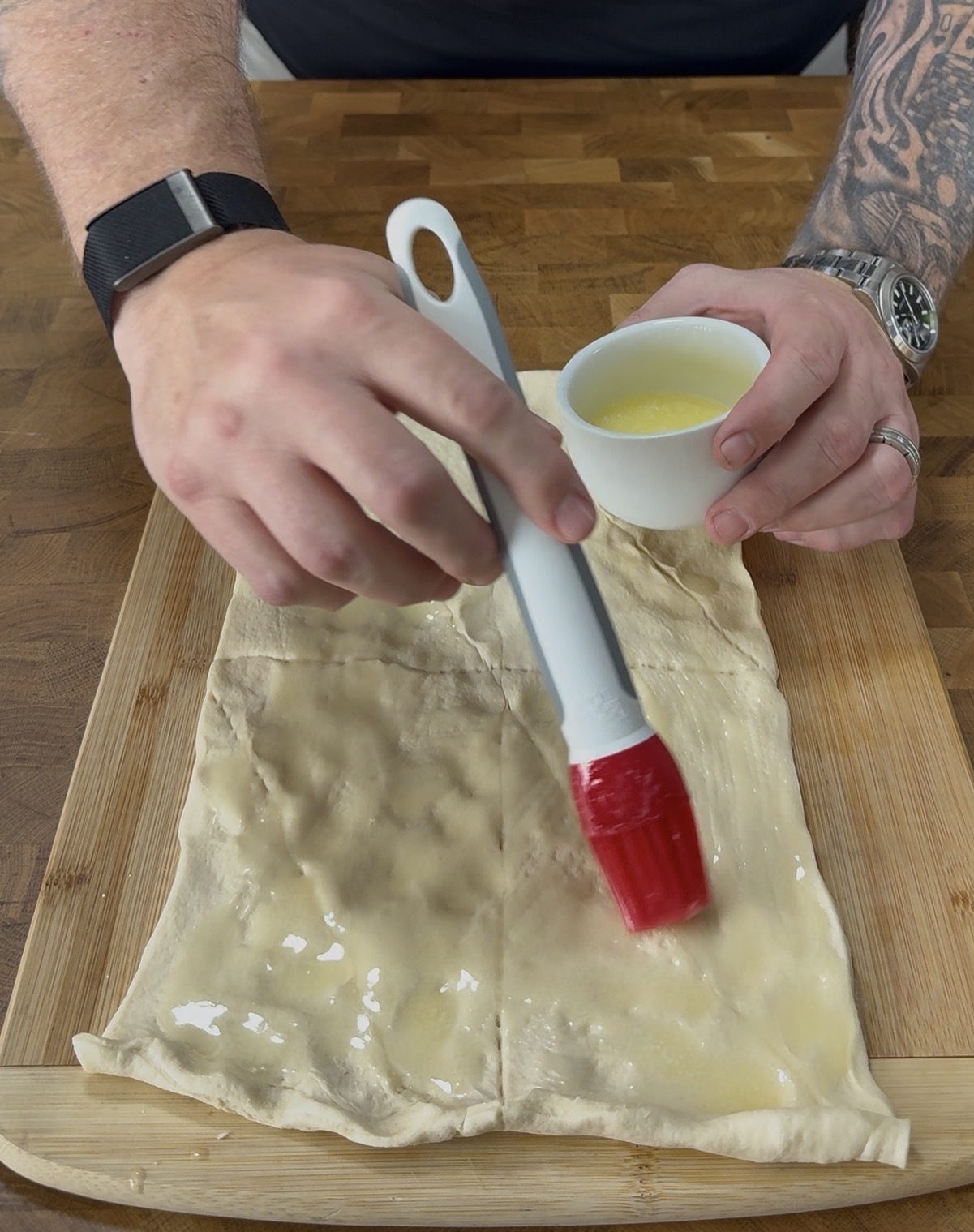 A person brushes melted butter onto a sheet of dough on a wooden board, holding a pastry brush in one hand and a small cup of butter in the other.