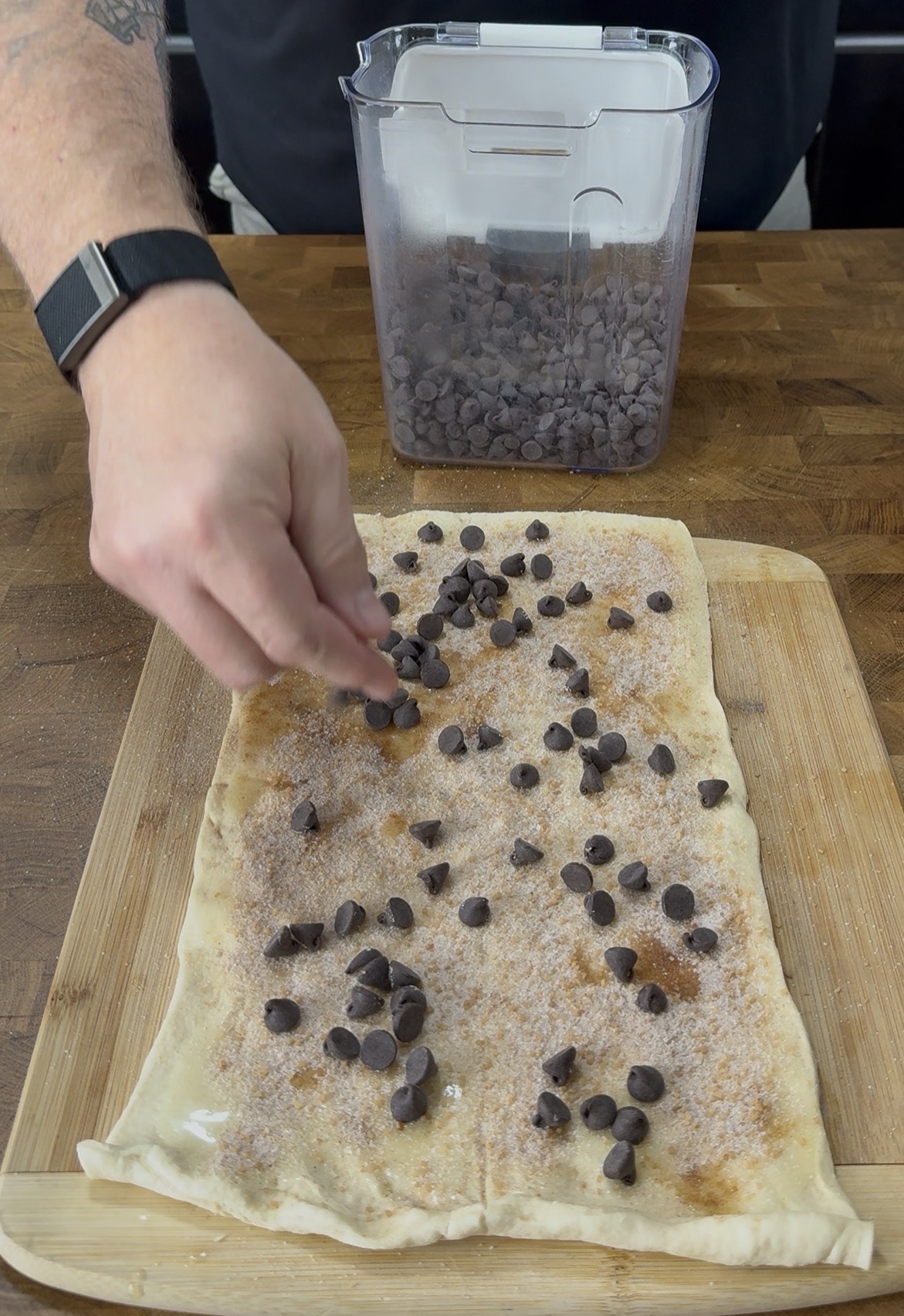 A hand sprinkles chocolate chips over a rectangular sheet of dough covered with cinnamon sugar, on a wooden cutting board. An open container of chocolate chips sits in the background.