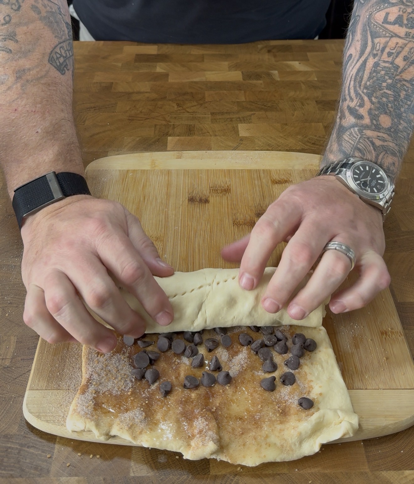A person with tattooed arms rolls dough topped with chocolate chips, sugar, and cinnamon on a wooden cutting board.
