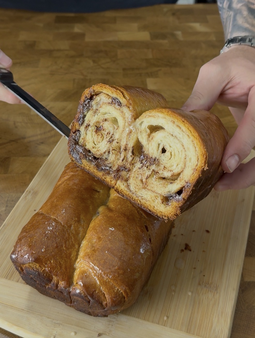 A person slices a loaf of swirled chocolate bread on a wooden cutting board, revealing the marbled layers inside.