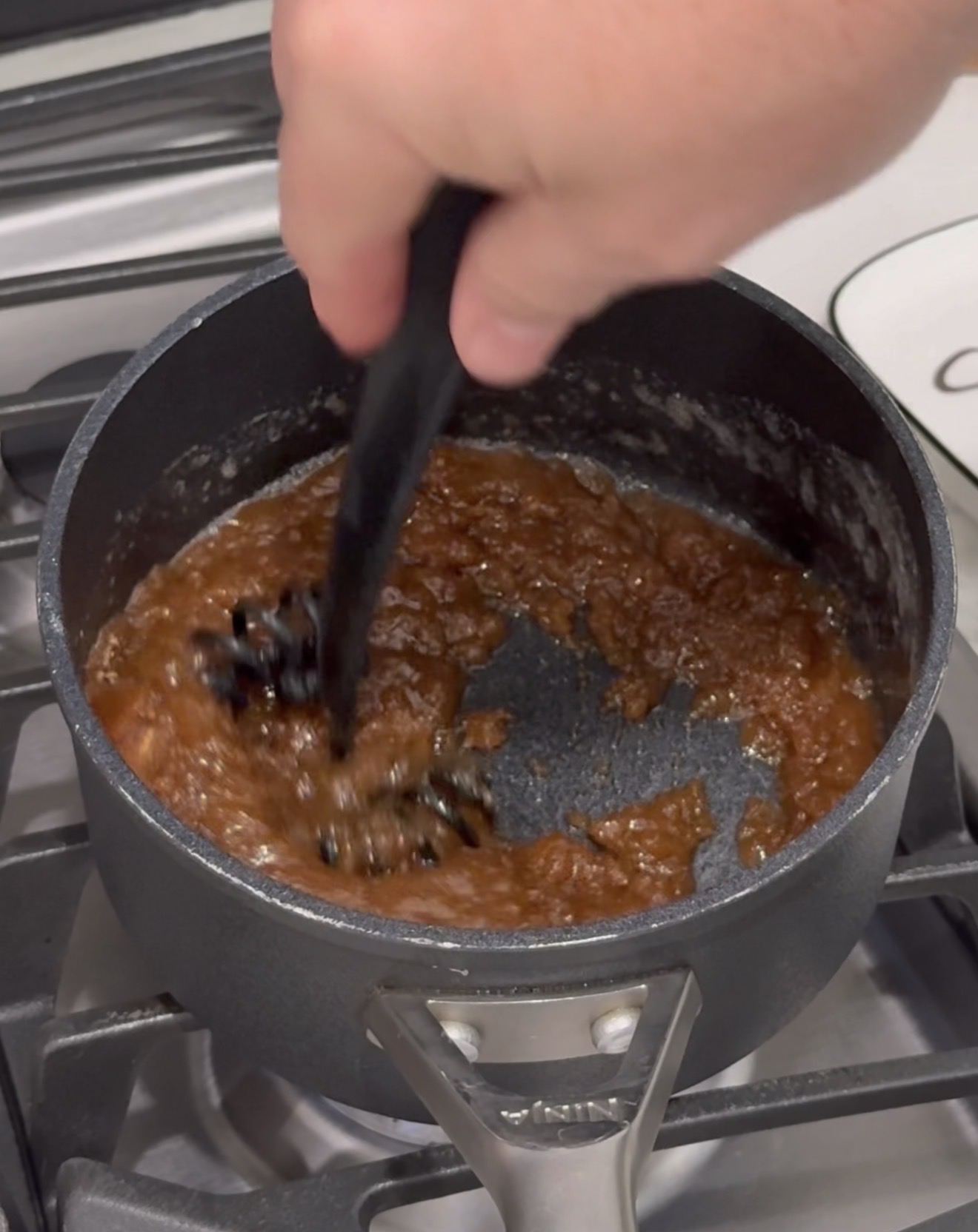A hand uses a black utensil to stir a thick, brown mixture in a small saucepan on a stovetop.