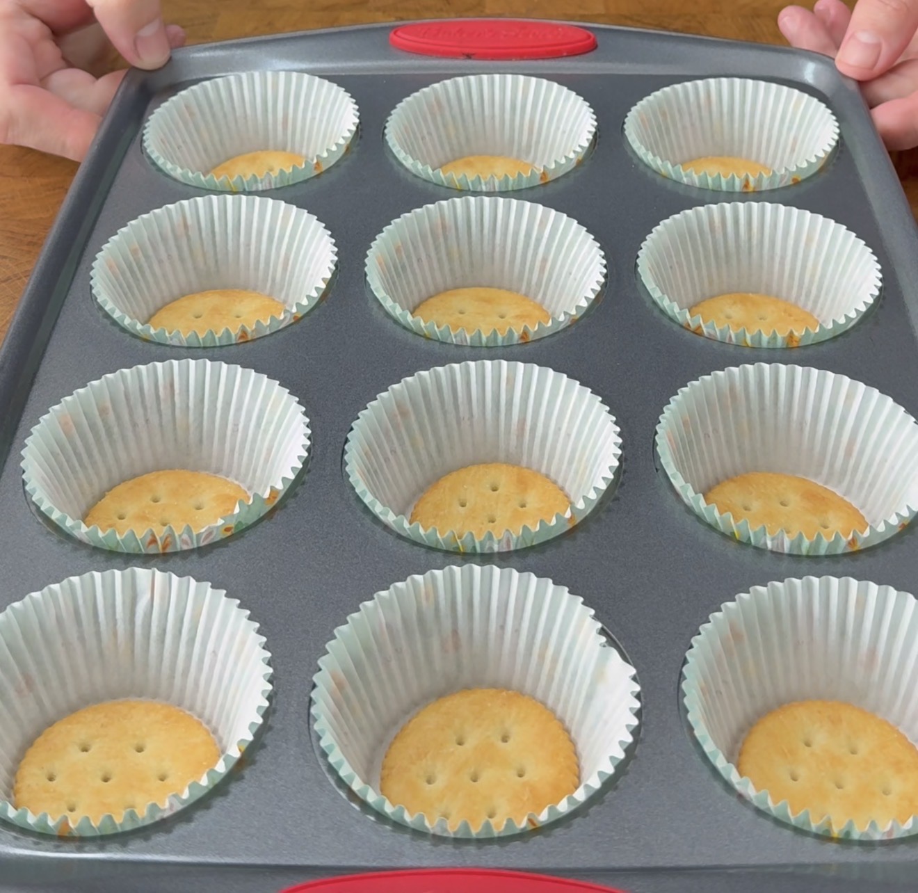 A muffin tin holds twelve paper liners, each with a round cracker at the bottom. Two hands are visible holding the sides of the tin.