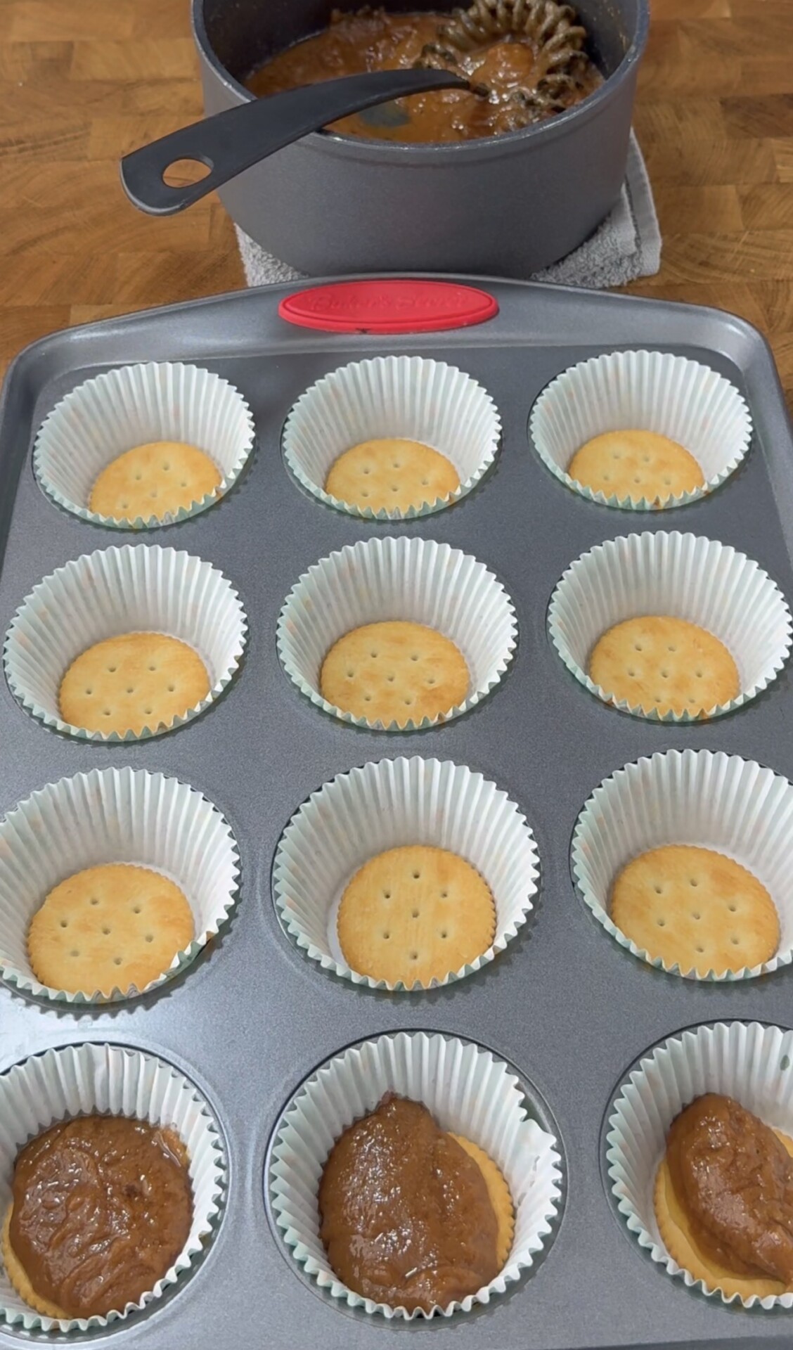 A muffin tray lined with paper cups, each holding a round cracker; some cups have a scoop of brown filling. In the background, a pot of brown mixture with a whisk sits on a counter.
