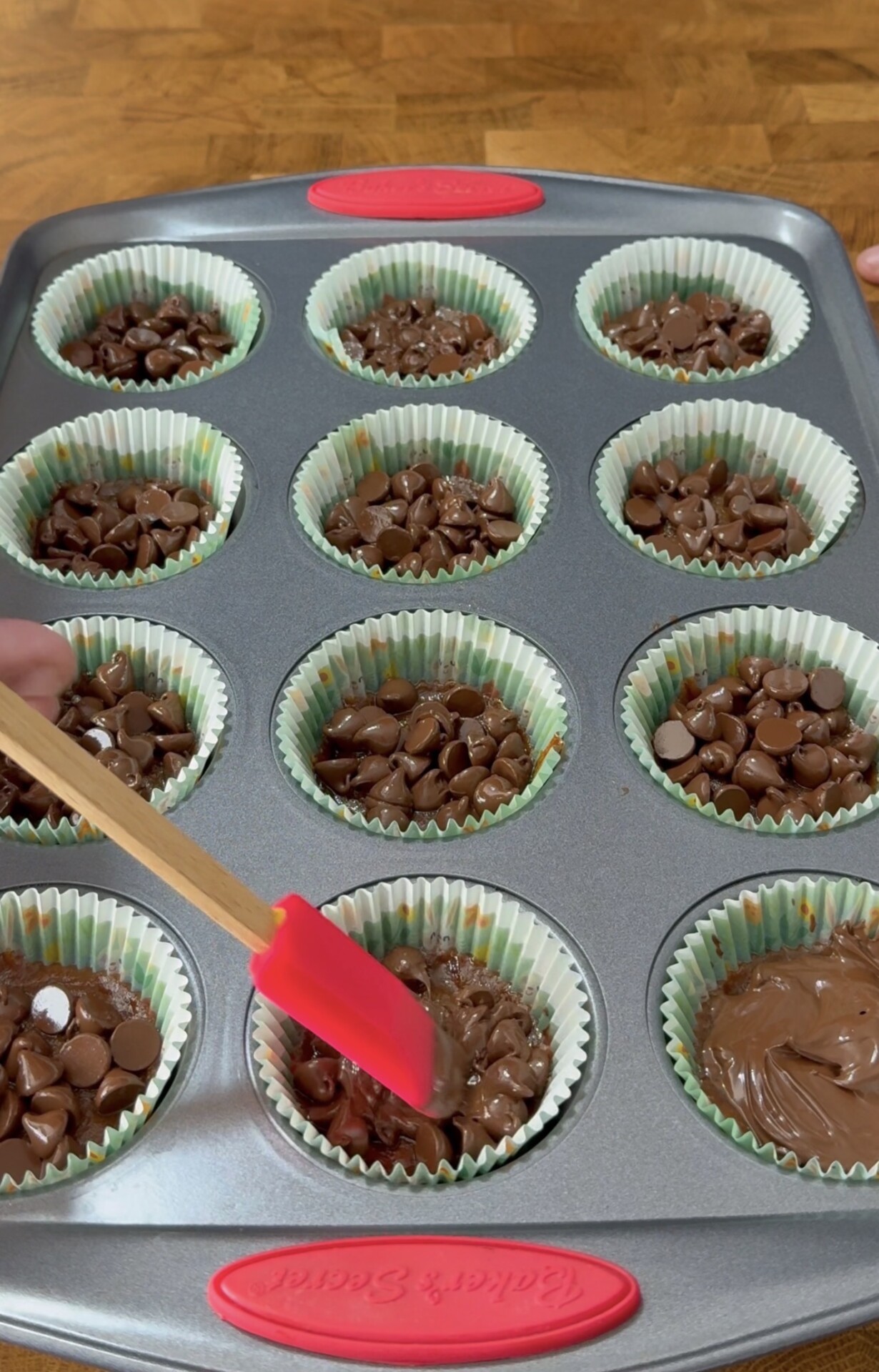 A muffin tin lined with paper cups is filled with chocolate batter and topped with chocolate chips. A hand holds a red spatula, spreading batter in one of the cups.