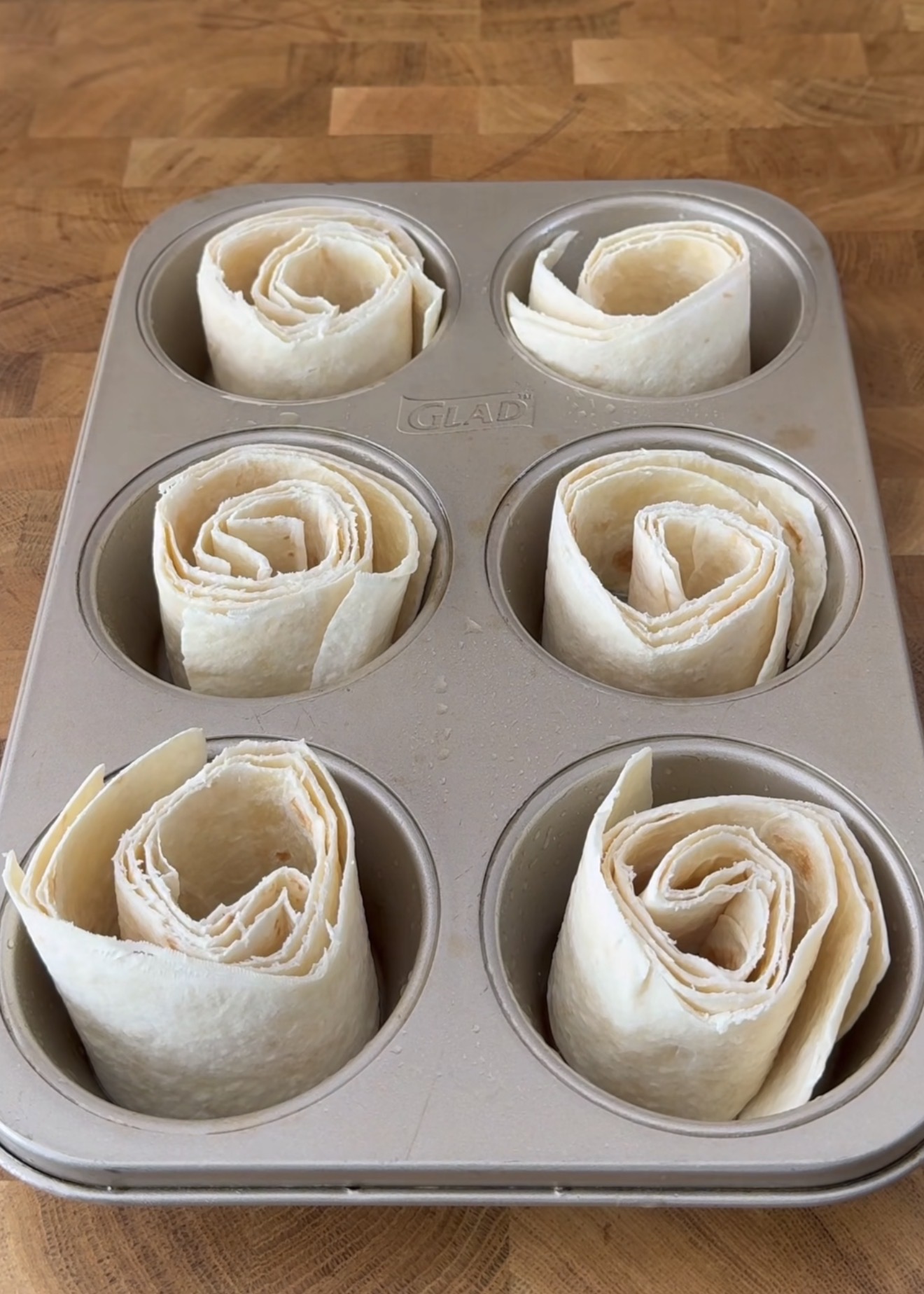 Six rolled flour tortillas are placed upright in the cups of a six-cup muffin tin, arranged in a spiral pattern. The tin is set on a wooden surface.