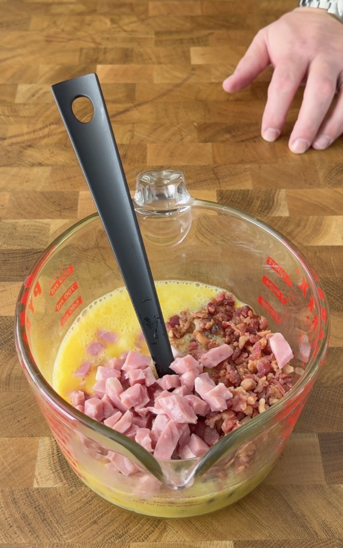 A measuring cup filled with beaten eggs, diced ham, and bacon bits sits on a wooden surface. A black spatula is in the cup, and a hand is visible in the background.