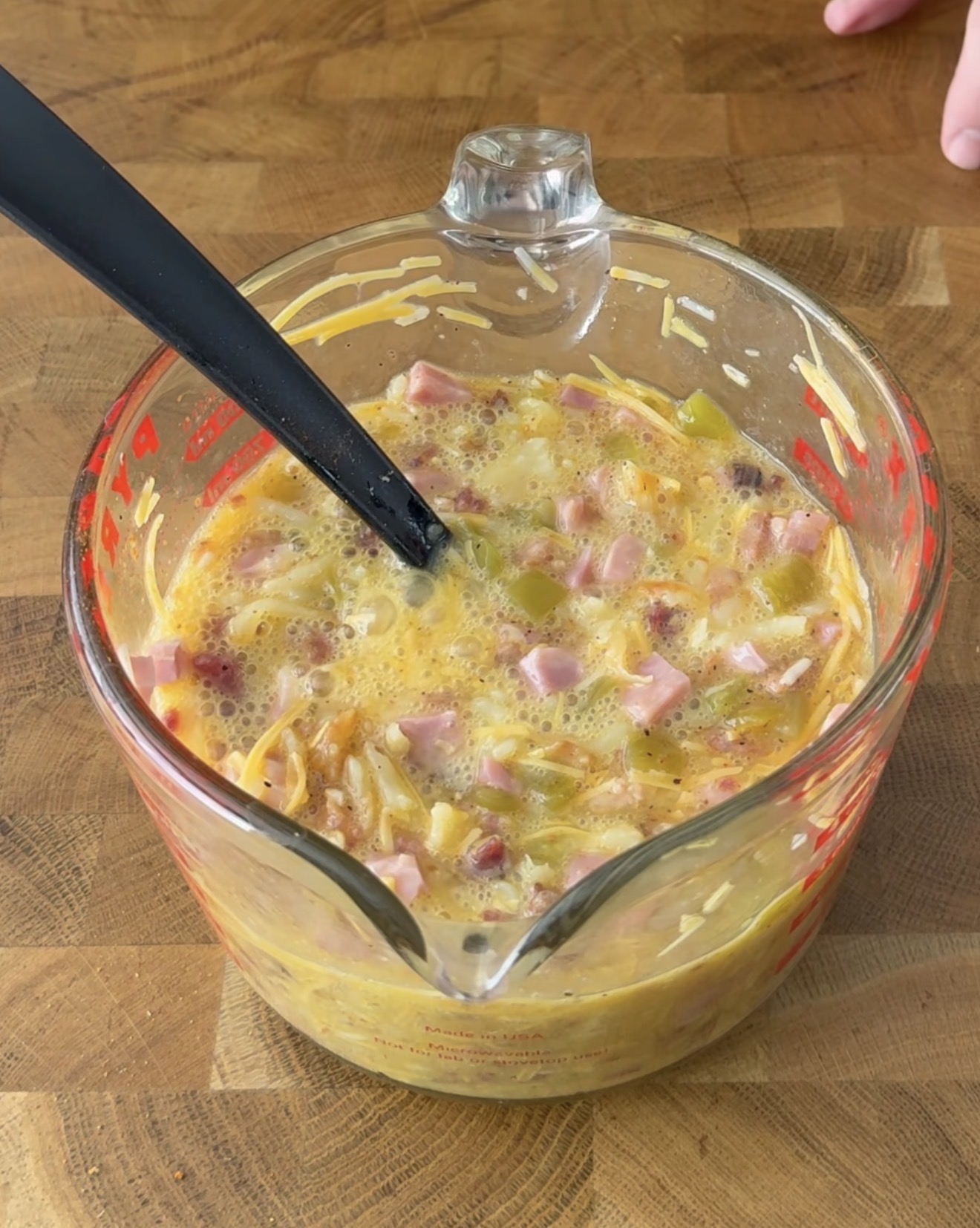 A glass measuring cup filled with a mixture of eggs, diced ham, shredded cheese, and chopped green bell peppers. A black spoon is resting inside the cup on a wooden surface. Two fingers are visible at the top right.