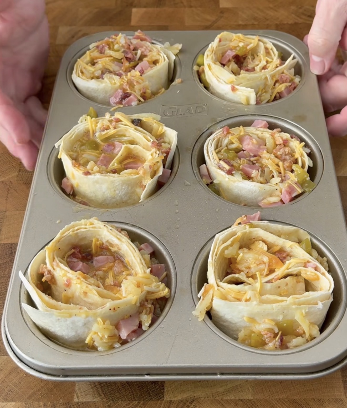 A muffin tin holds six unbaked tortilla cups filled with a mixture of chopped ham, shredded cheese, and vegetables on a wooden surface. Hands are positioned near the tin, ready to move or adjust it.