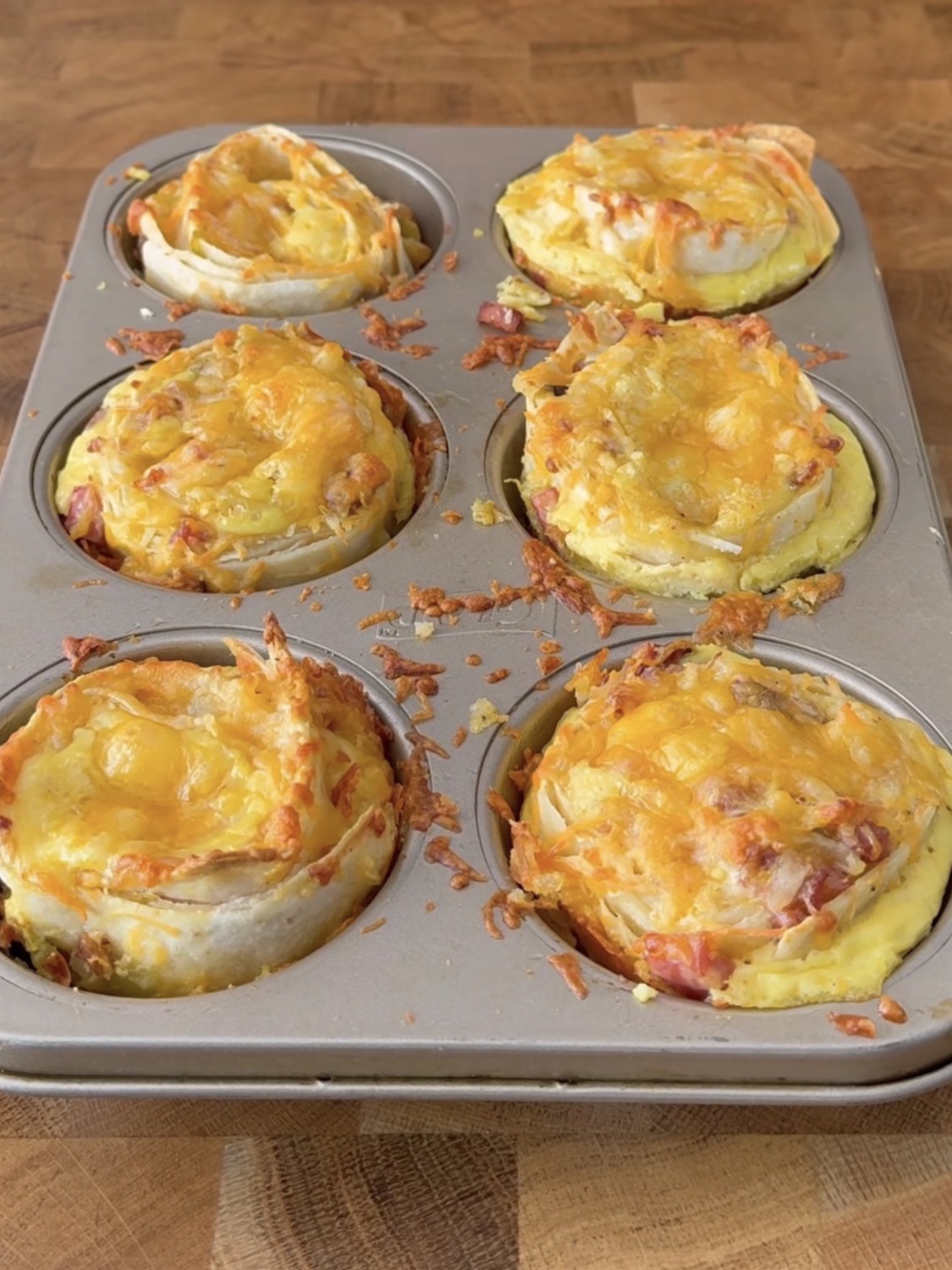 A muffin tin filled with six baked egg and cheese breakfast cups, each topped with melted cheese and bits of ham, resting on a wooden surface.