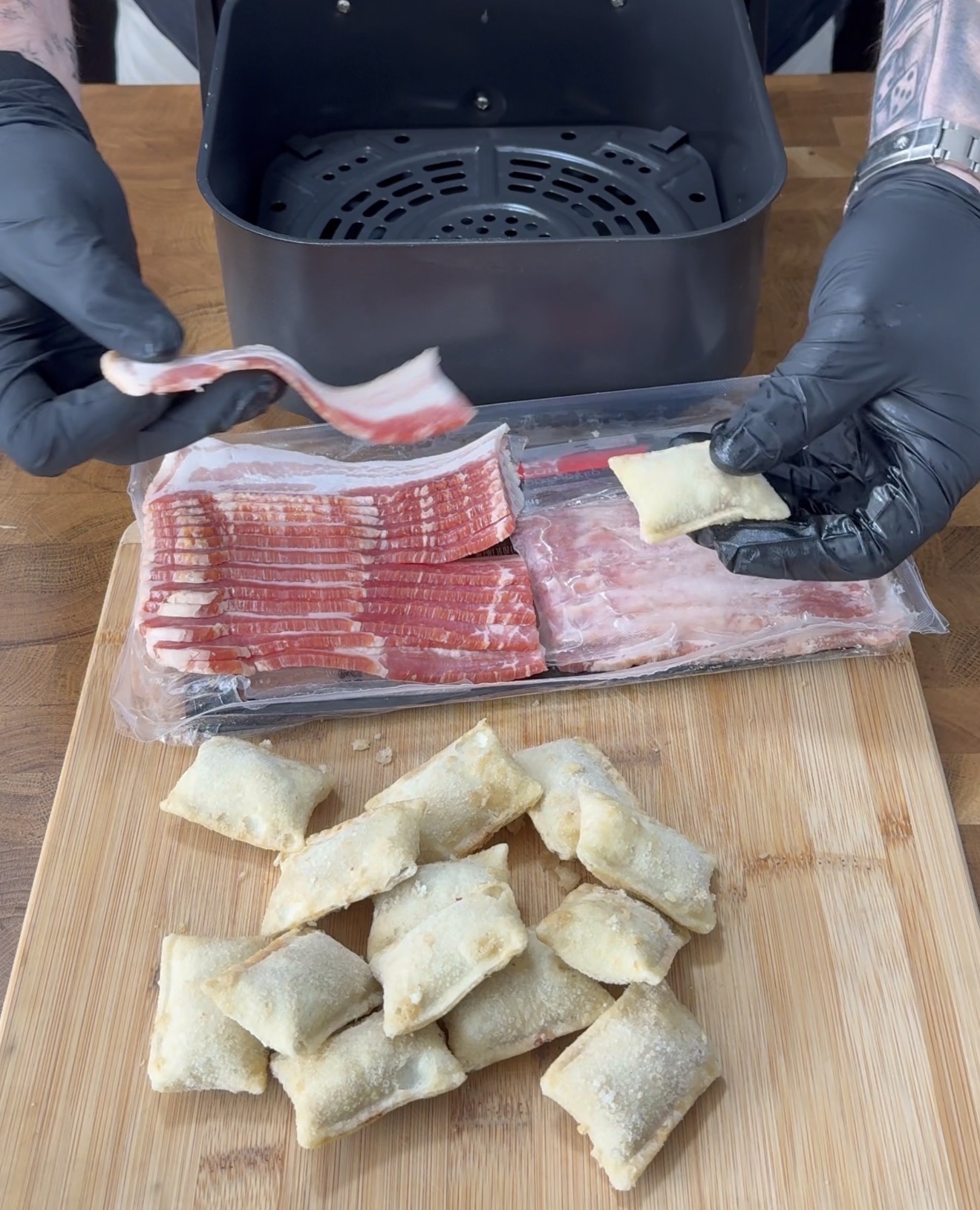 A person wearing black gloves wraps a frozen pizza roll with a strip of raw bacon in front of an air fryer. Uncooked bacon strips and several frozen pizza rolls rest on a wooden cutting board.
