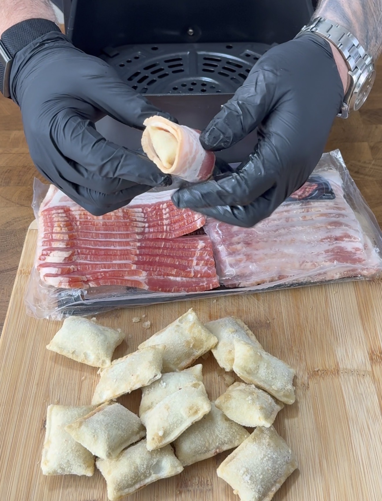 A person wearing black gloves wraps a pizza roll with a slice of bacon over a wooden board. Frozen pizza rolls and a pack of uncooked bacon are visible in the background.