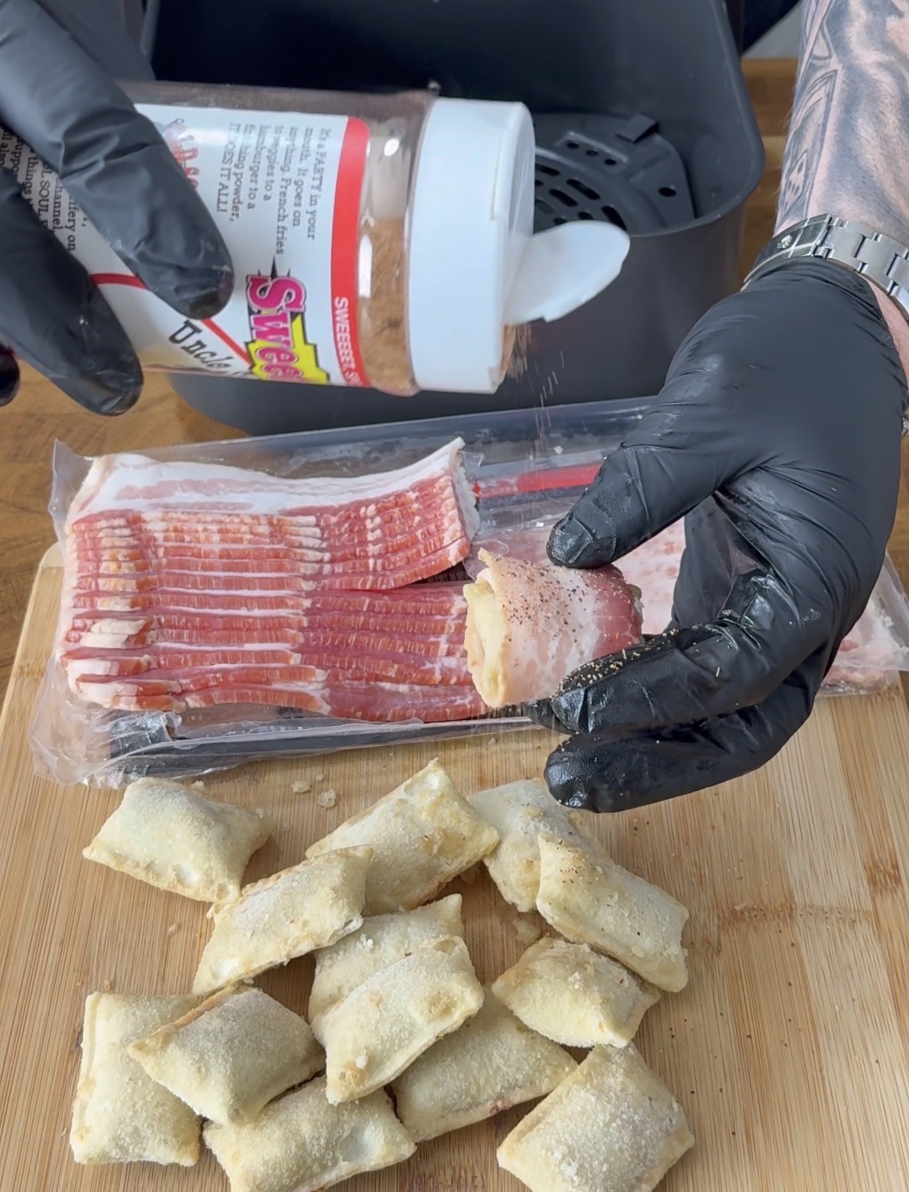A person wearing black gloves sprinkles seasoning onto a pizza roll wrapped in bacon over a wooden cutting board, with more pizza rolls, bacon slices, and an air fryer visible in the background.