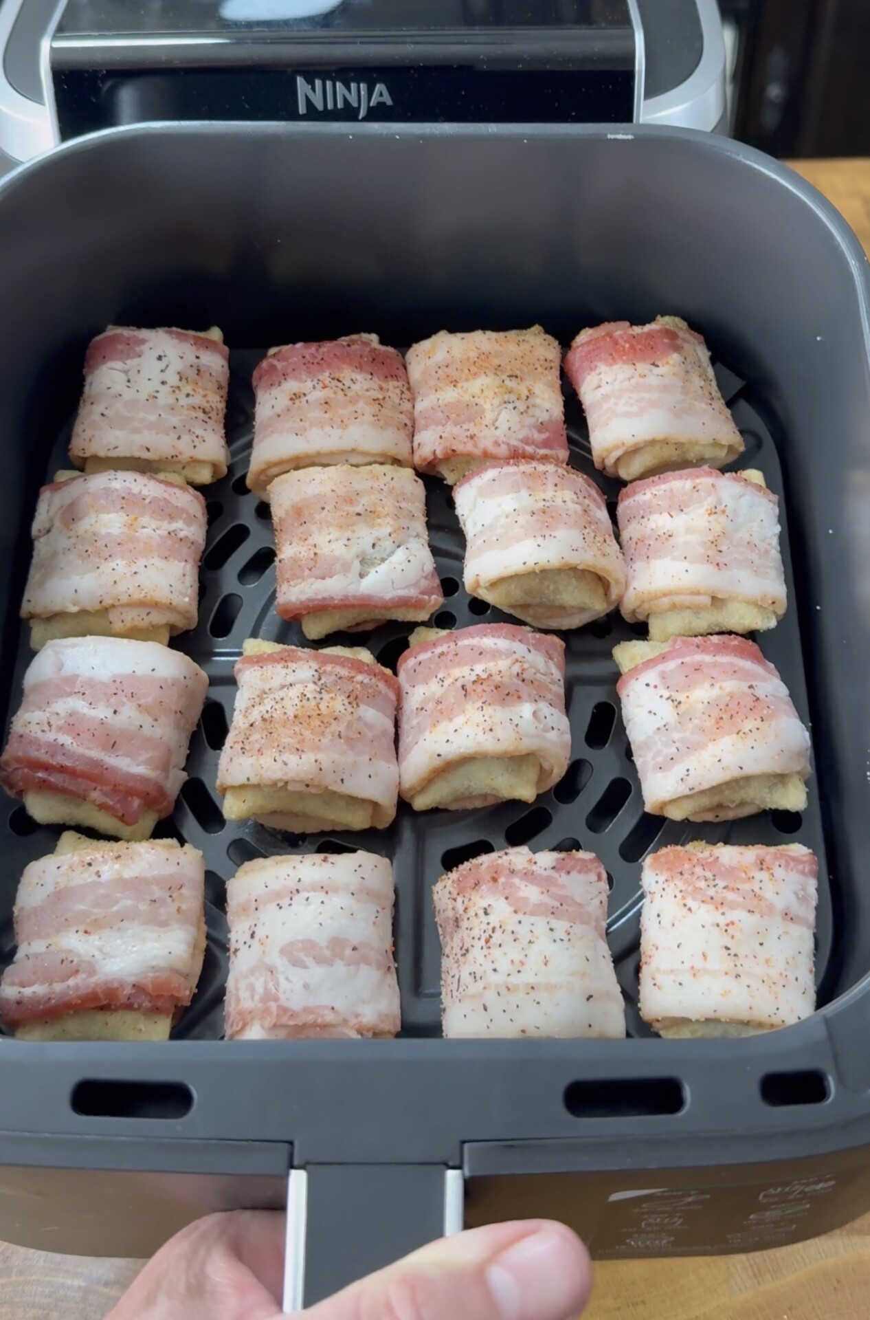 Raw bacon-wrapped pieces of food arranged in a single layer inside an air fryer basket, ready to be cooked. A hand is holding the air fryer drawer, and the appliance brand “Ninja” is visible at the top.