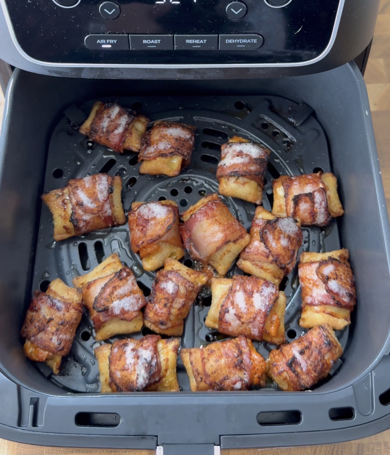 Bacon-wrapped snacks are arranged in a single layer inside an air fryer basket. The snacks appear cooked, with the bacon crispy and browned. The air fryer control panel is partially visible at the top of the image.