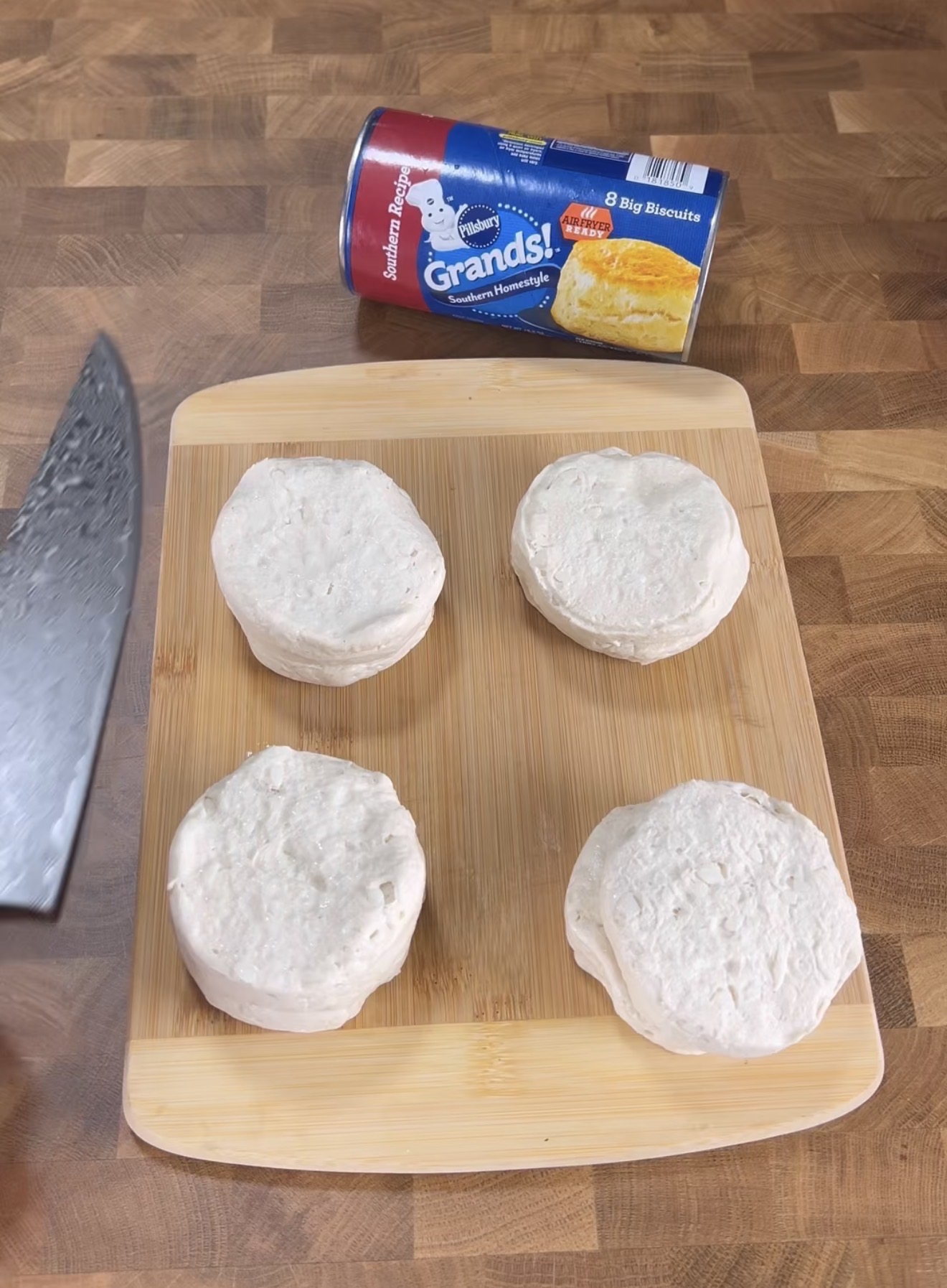 Four raw biscuit dough rounds are arranged on a wooden cutting board. An open can of Pillsbury Grands! Southern Homestyle biscuits is above the board, and a knife is partially visible on the left side.