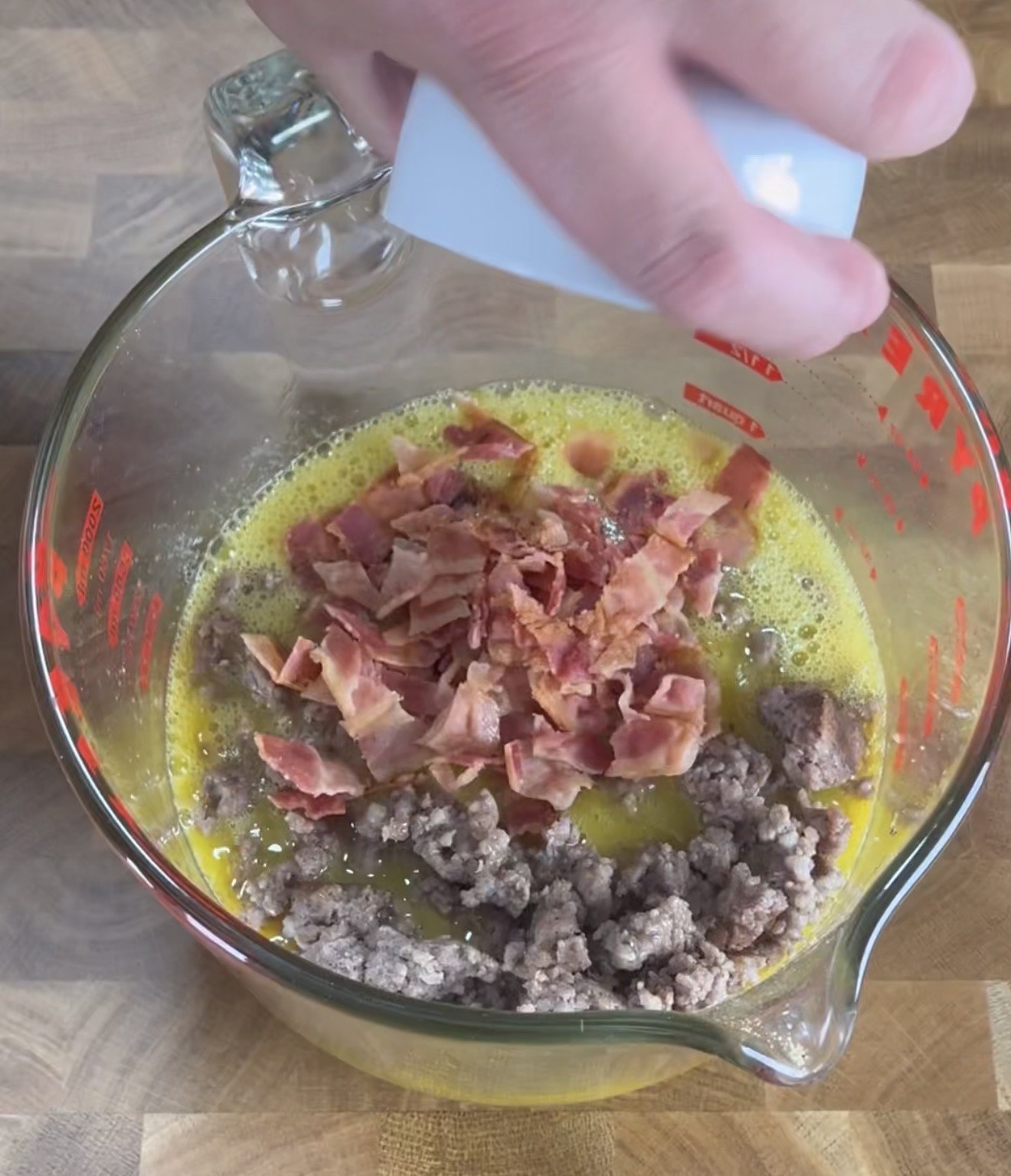 A hand holds a white cup above a glass measuring bowl filled with beaten eggs, crumbled cooked bacon, and cooked ground sausage, sitting on a wooden surface.