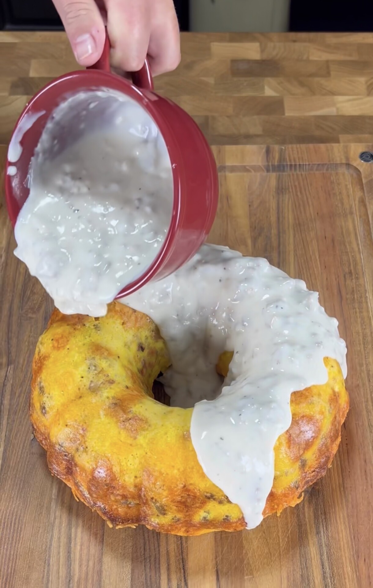A hand pours thick white gravy from a red cup onto a golden-brown Bundt-shaped dish on a wooden cutting board.