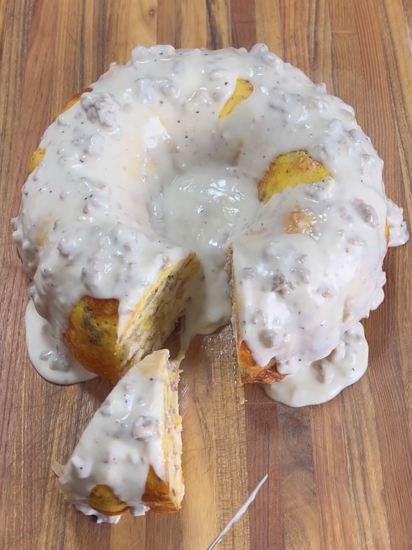 A bundt-shaped bread covered in creamy sausage gravy sits on a wooden surface, with one slice cut and slightly pulled out from the loaf.
