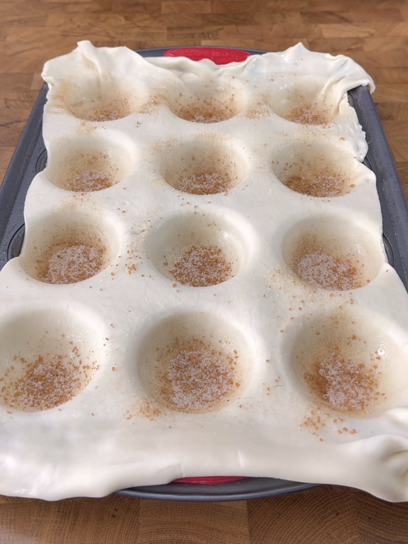A sheet of dough is draped over an inverted muffin tin, with indentations sprinkled with cinnamon and sugar, on a wooden surface.
