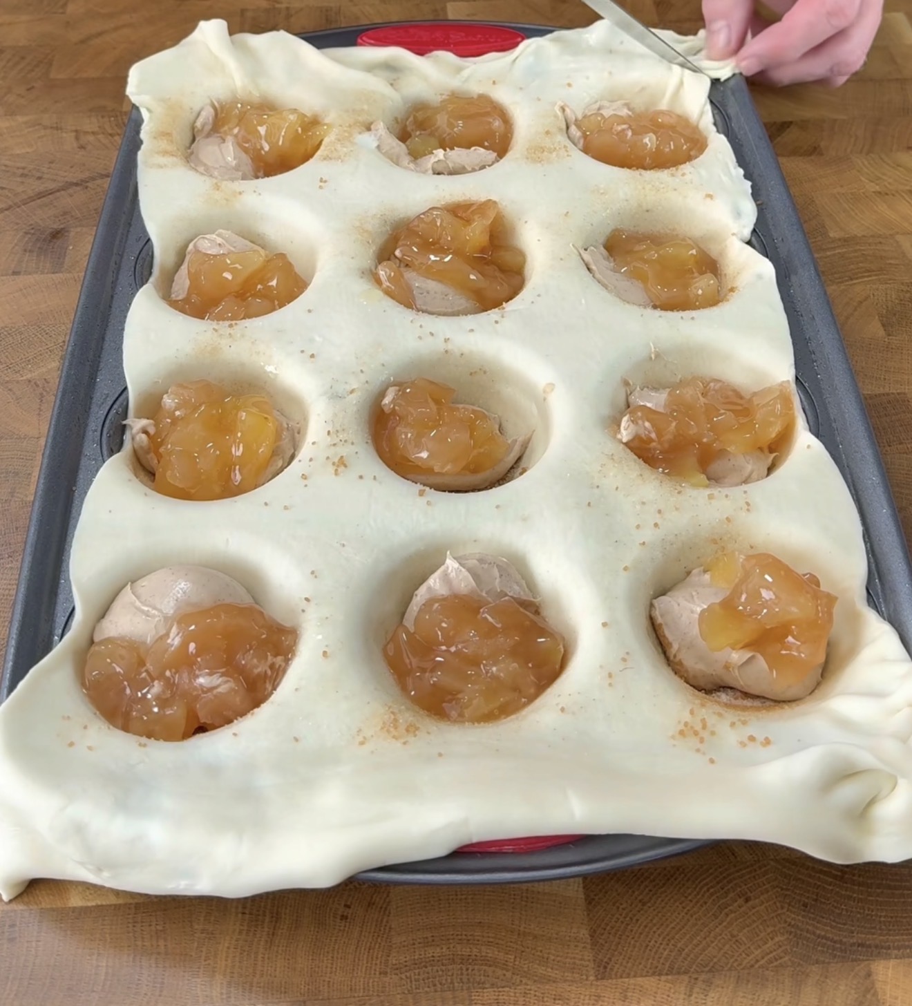 A baking tray holds a sheet of dough pressed into wells, each filled with apple pie filling. A hand uses a knife to cut excess dough along the tray edge on a wooden countertop.