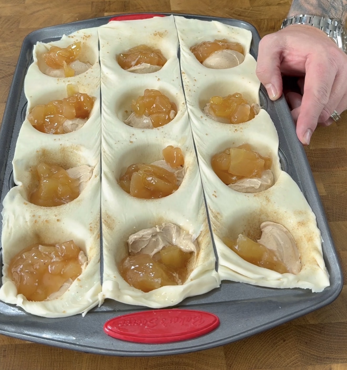 A muffin tin lined with dough is filled with scoops of apple pie filling and creamy mixture. A person’s hand is holding the pan on a wooden counter.