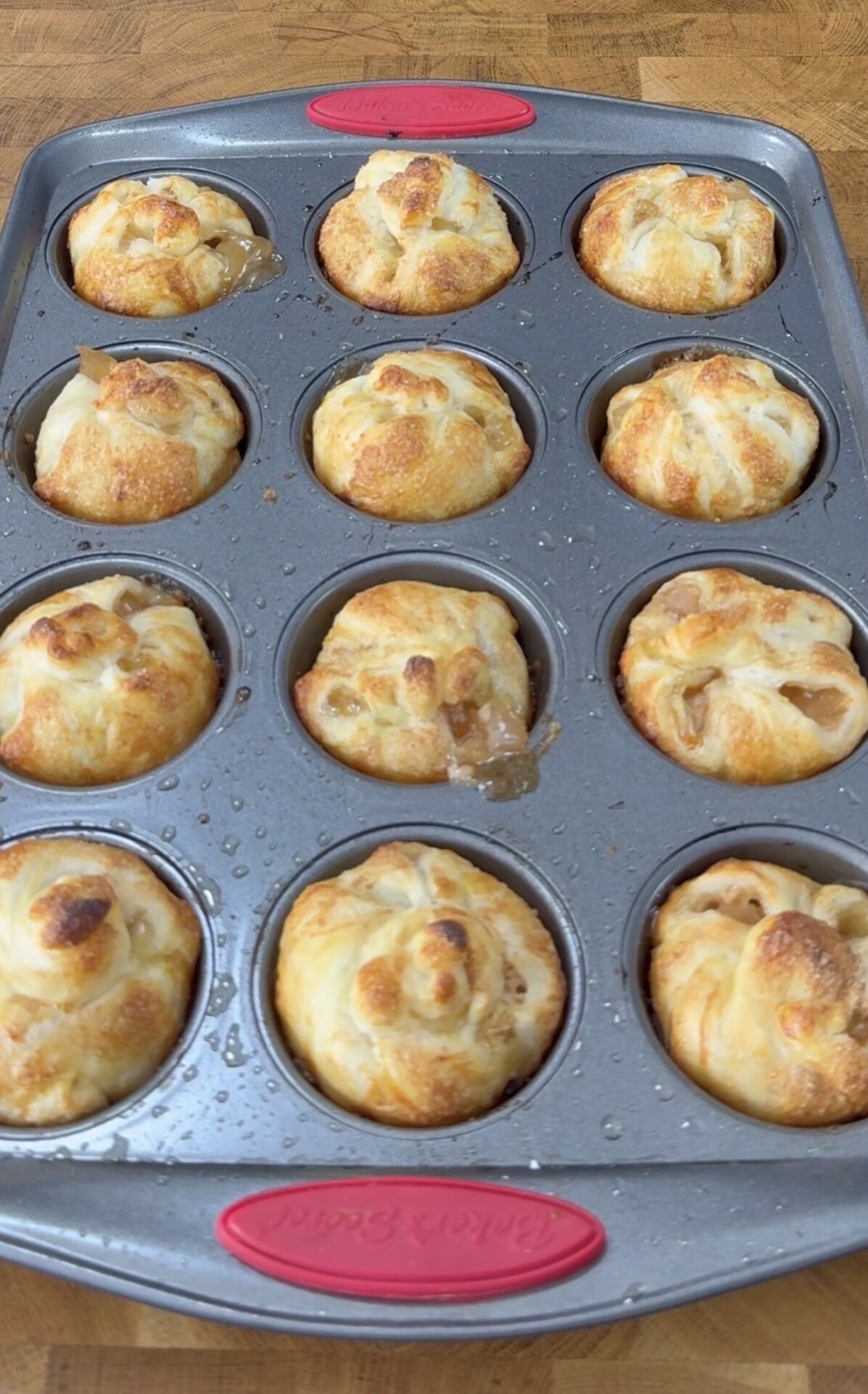 A muffin tin holds twelve golden-brown, puff pastry cups, some with filling visible and slightly oozing out. The pan has red silicone grips and sits on a wooden surface.
