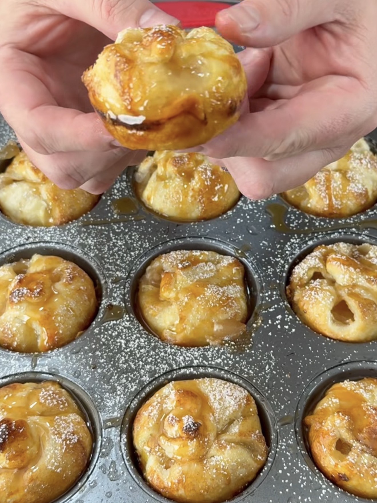 A person holds a small pastry drizzled with glaze above a muffin tin filled with baked pastries, each dusted with powdered sugar.