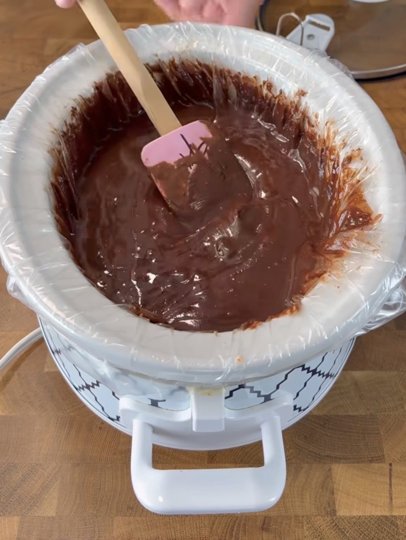 A crockpot lined with a plastic liner is filled with melted chocolate being stirred with a pink spatula on a wooden countertop.