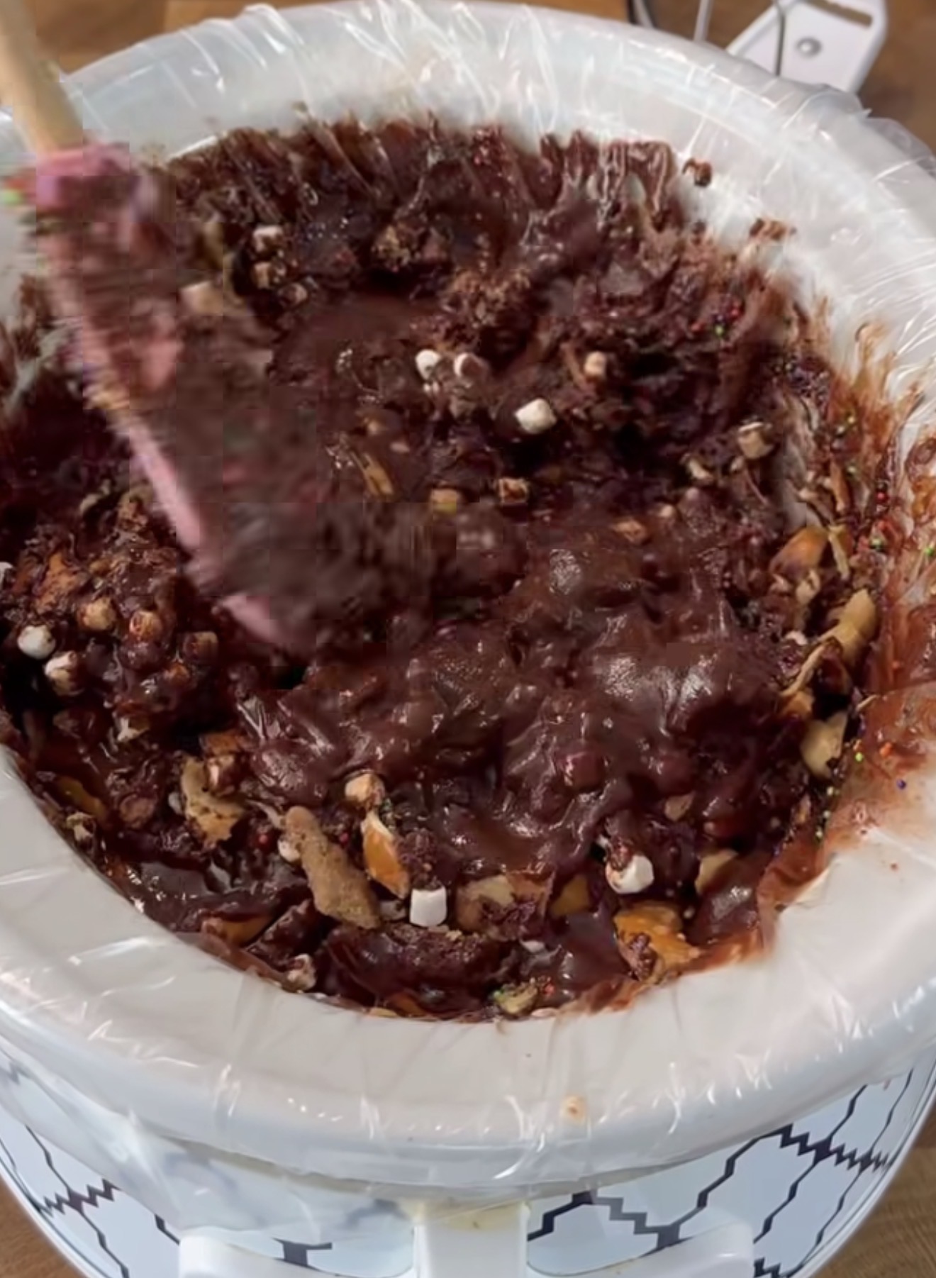 A close-up of a crockpot lined with a plastic bag, filled with melted chocolate, nuts, and marshmallows being stirred with a pink spatula.