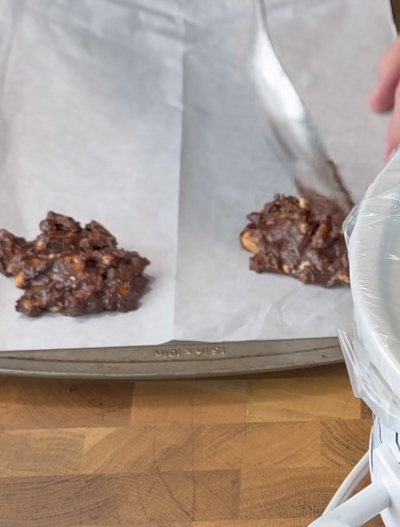 Two scoops of chocolate cookie dough are placed on a parchment-lined baking sheet, with a hand using a spoon to shape one of the mounds. A mixing bowl covered in plastic wrap is partially visible in the foreground.