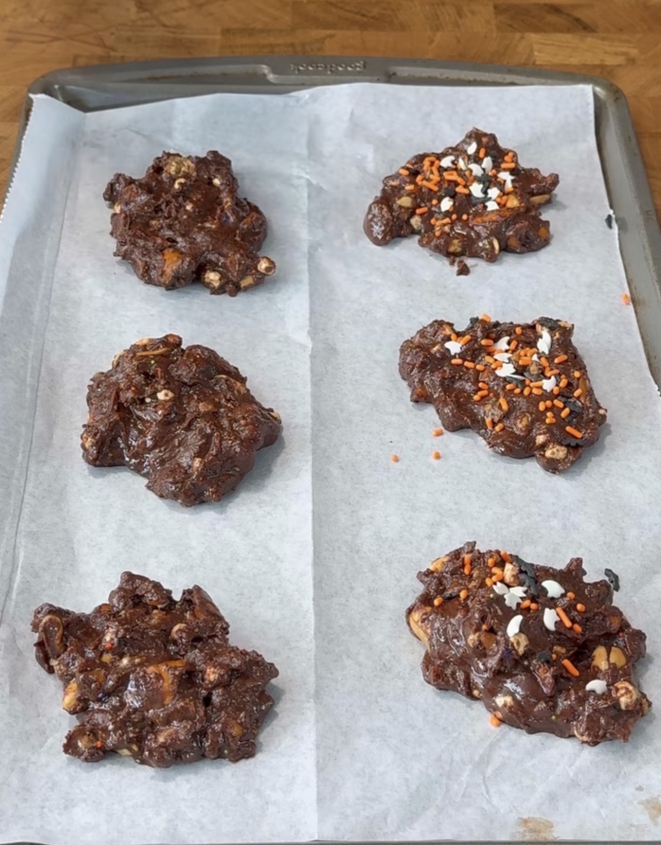 Six unbaked chocolate cookie dough mounds on a parchment-lined baking tray, with some topped with orange, white, and black sprinkles.