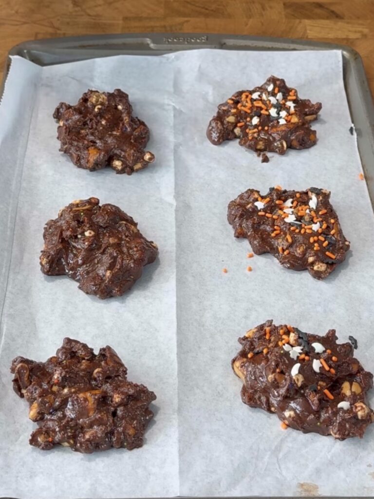 Six unbaked chocolate cookies with chunks are arranged on a parchment-lined baking sheet. Half have orange and white sprinkles on top, while the others do not. The baking sheet is on a wooden surface.